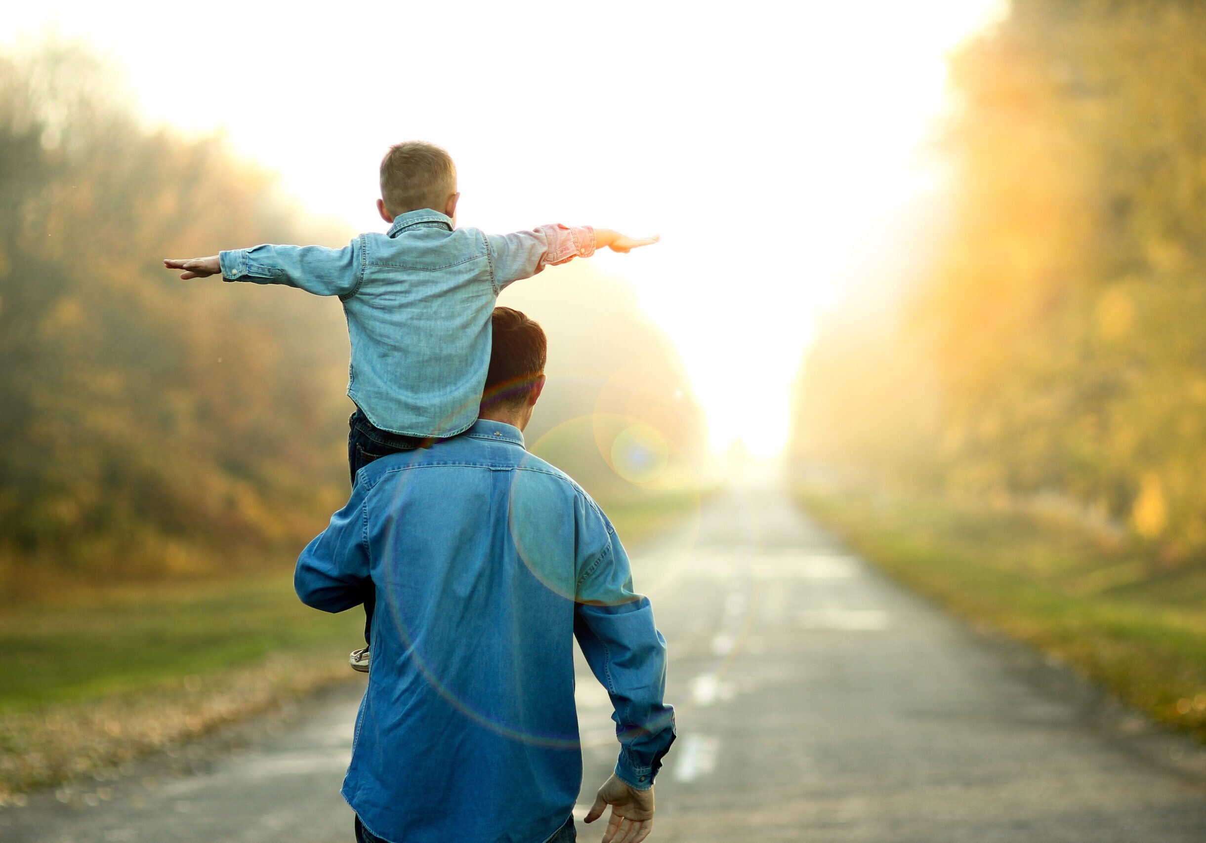 Father carrying child on his shoulders while walking down a sunlit country road.