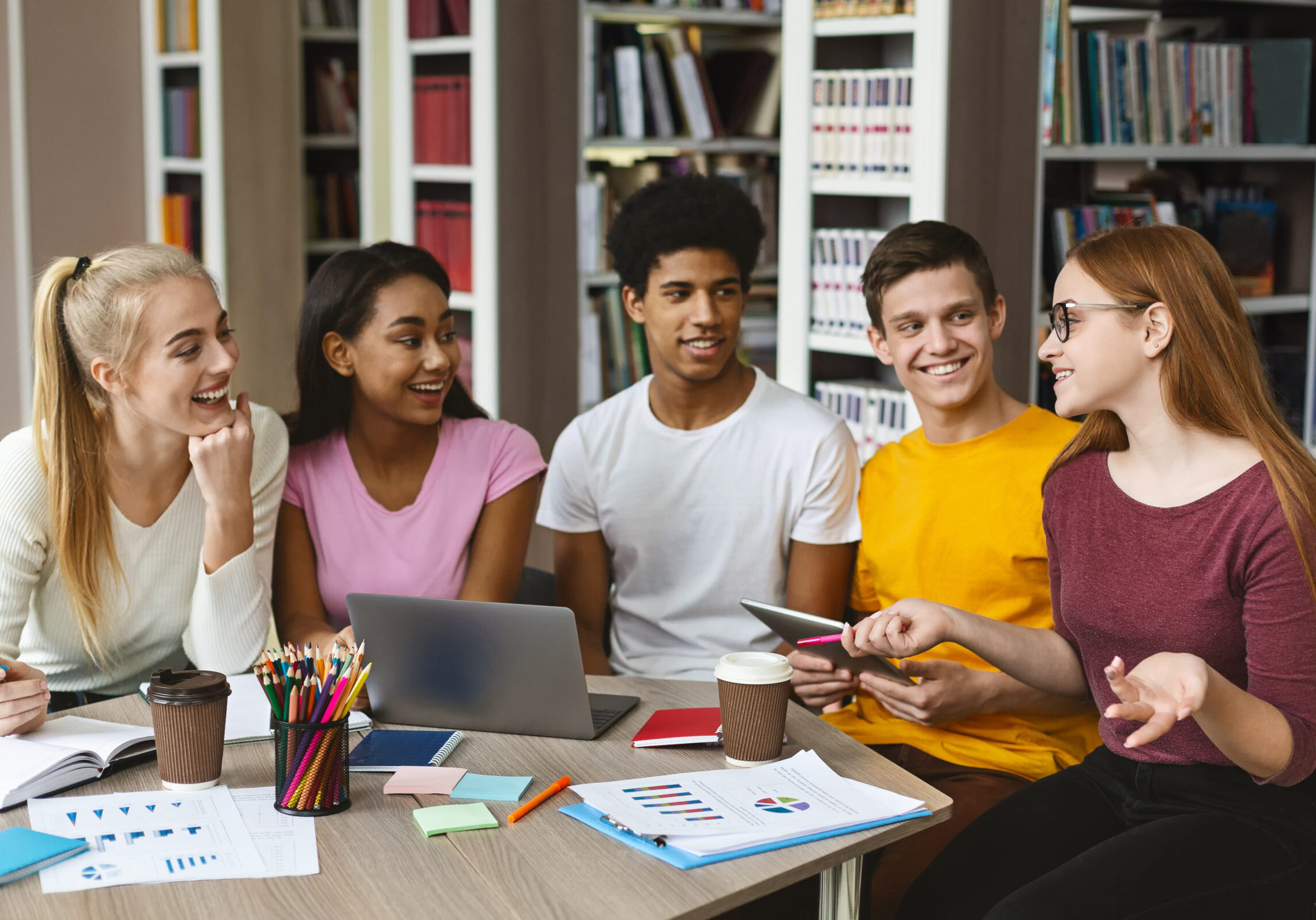 Group of diverse students collaborating around a table with laptops, charts, and notebooks in a library setting